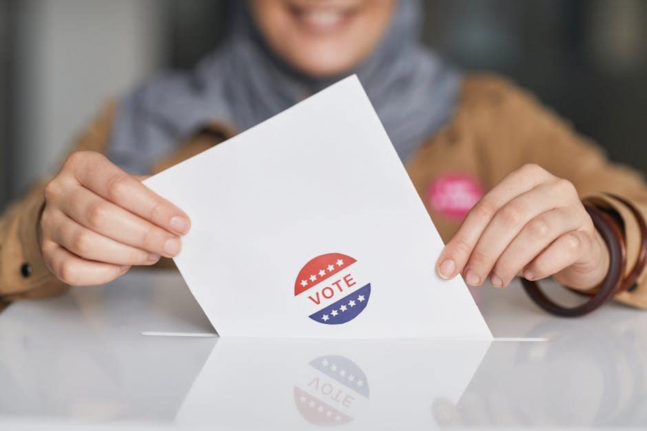 A person wearing a wristband fills out a voting ballot with a pen. Voting stickers visible.