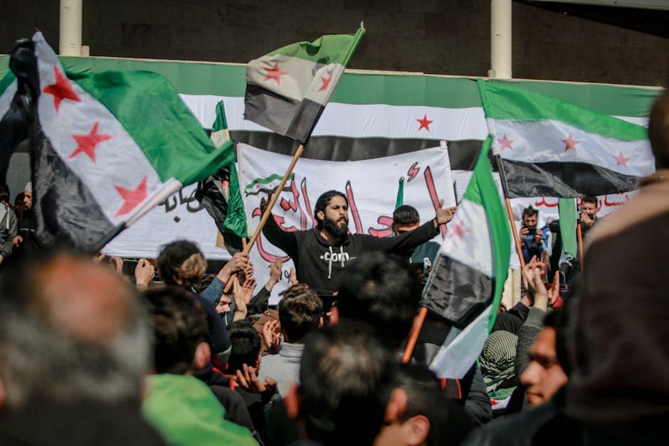 Protestors gather in Idlib, Syria, waving Syrian flags during a daytime demonstration.