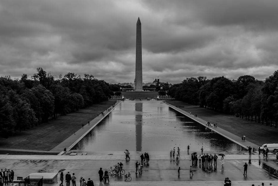 The Washington Monument stands tall reflected in the wet plaza after rain on a cloudy day.