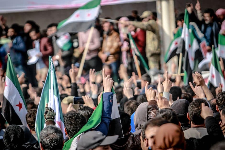 Crowds gather in Idlib, Syria, displaying national flags and banners during a protest.