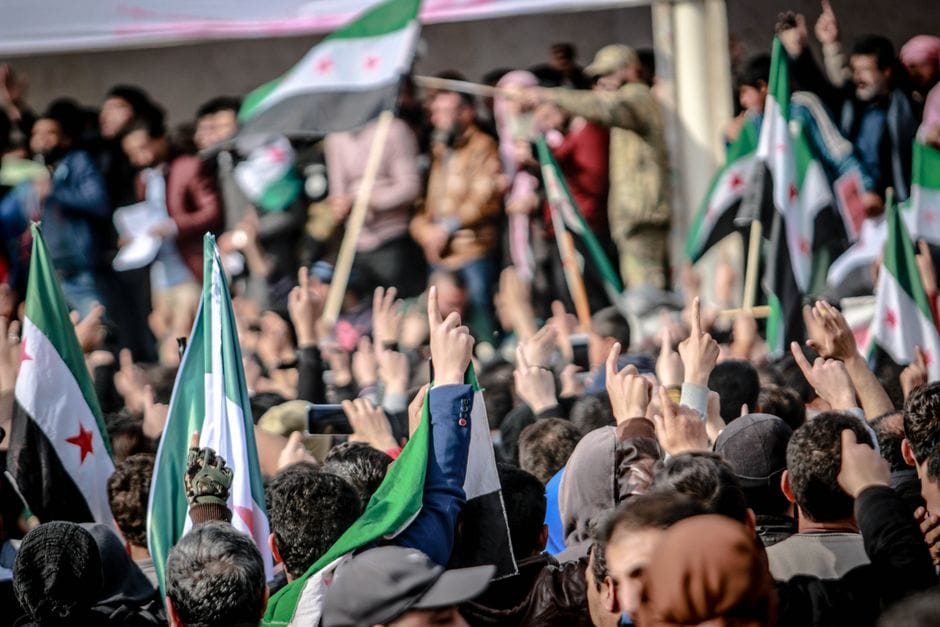 Crowds gather in Idlib, Syria, displaying national flags and banners during a protest.