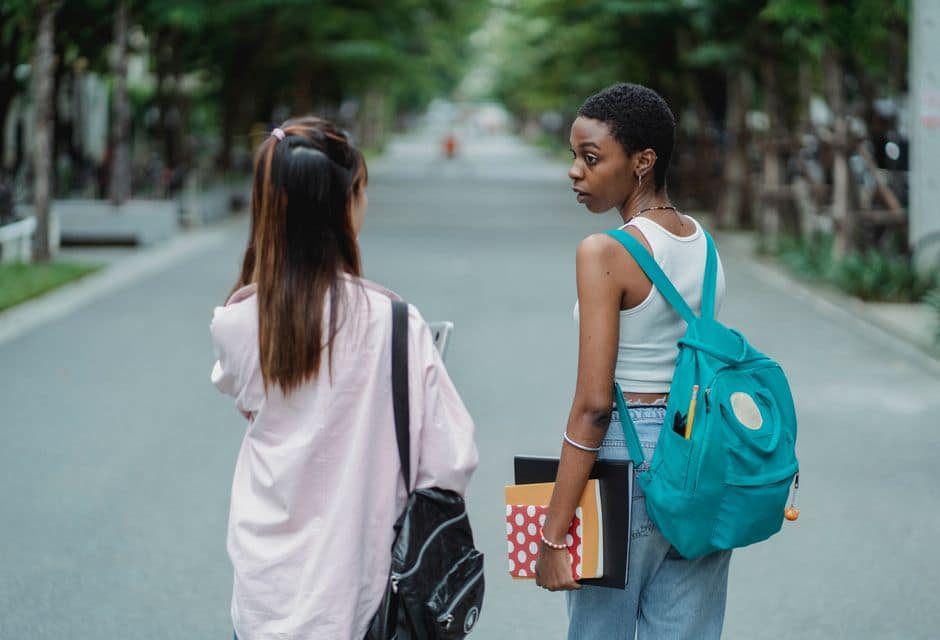 Back view of diverse female students carrying backpacks and supplies for university while talking in park