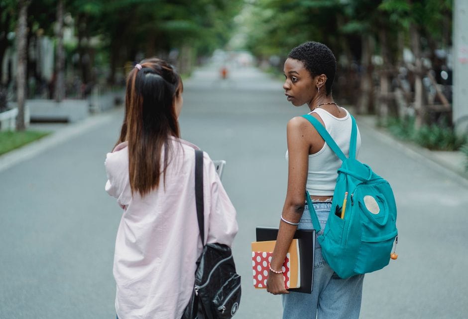 Back view of diverse female students carrying backpacks and supplies for university while talking in park