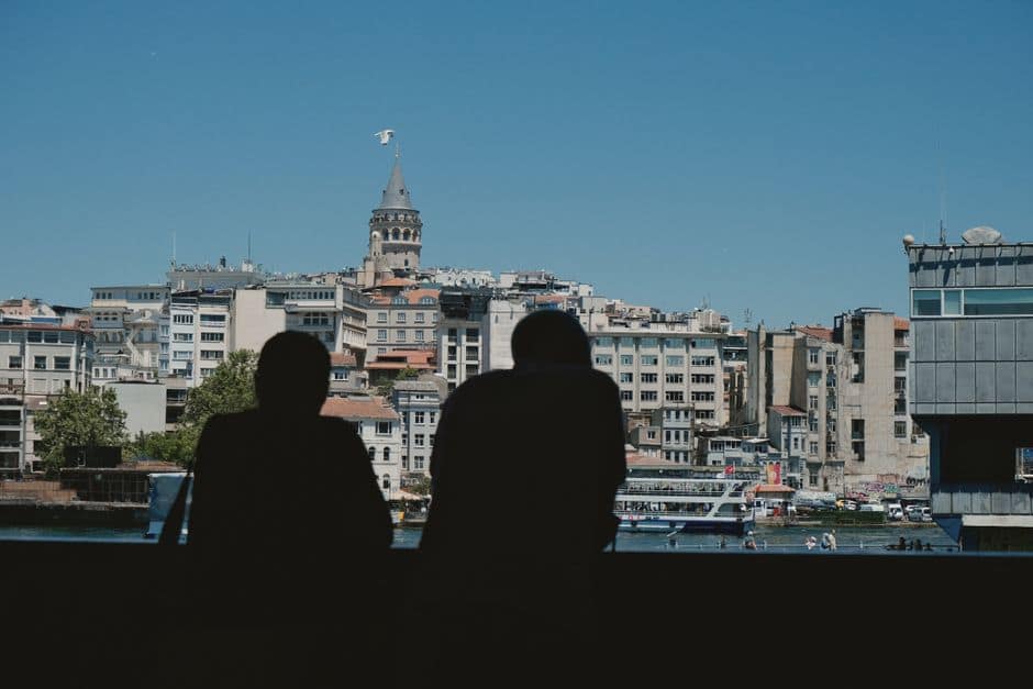 Two silhouettes view the Galata Tower and skyline from across the water on a sunny day in İstanbul.