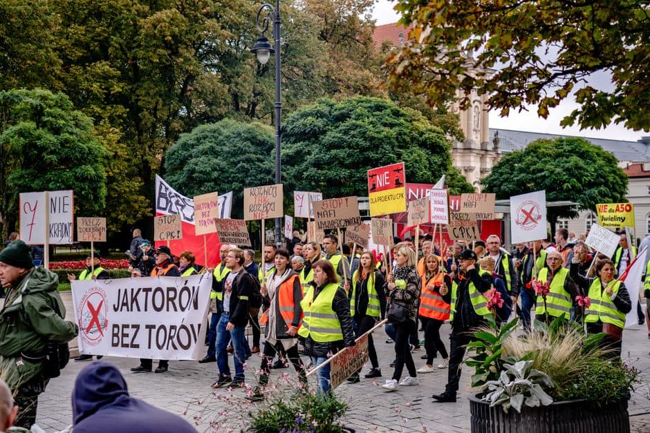A large group protests in Warsaw's city center, holding placards with messages in Polish.