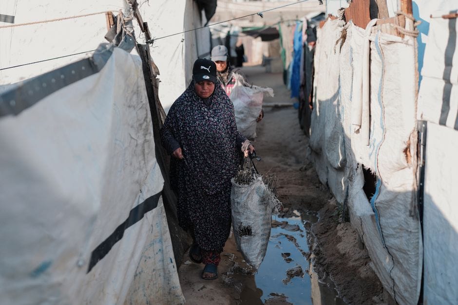 Two women navigate a narrow alley in a refugee camp, carrying bags. Real-life struggle captured.