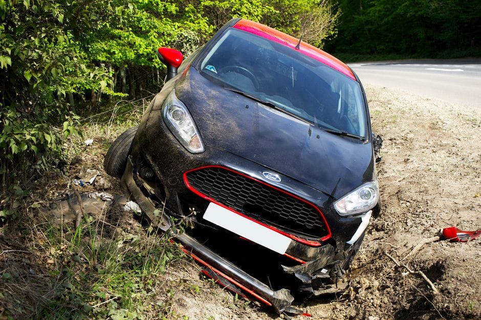 A black car, seemingly a Ford, crashed and damaged in a roadside ditch with surrounding greenery.