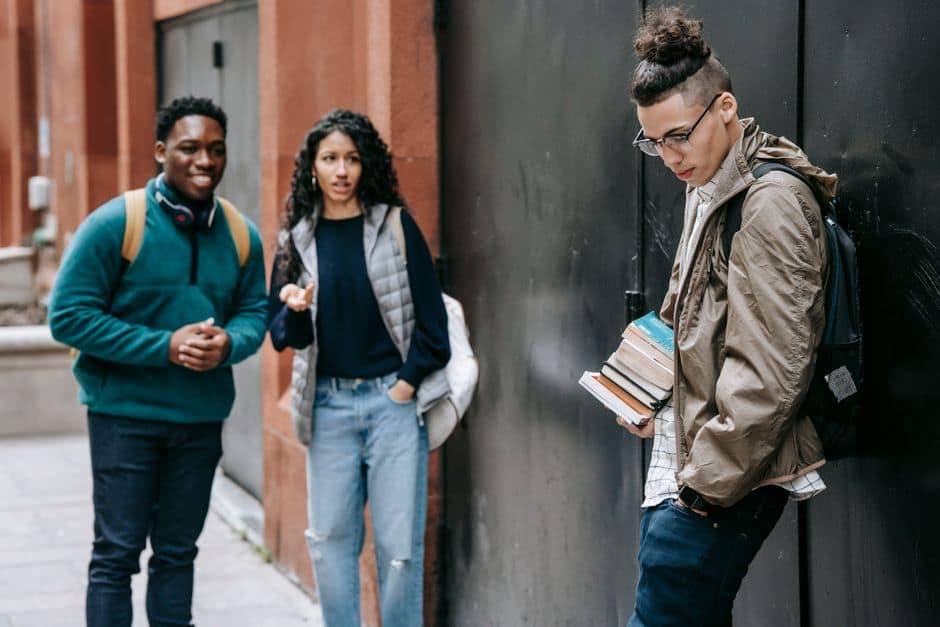 Cheerful multiracial students with backpacks standing near university building and bullying friend with books