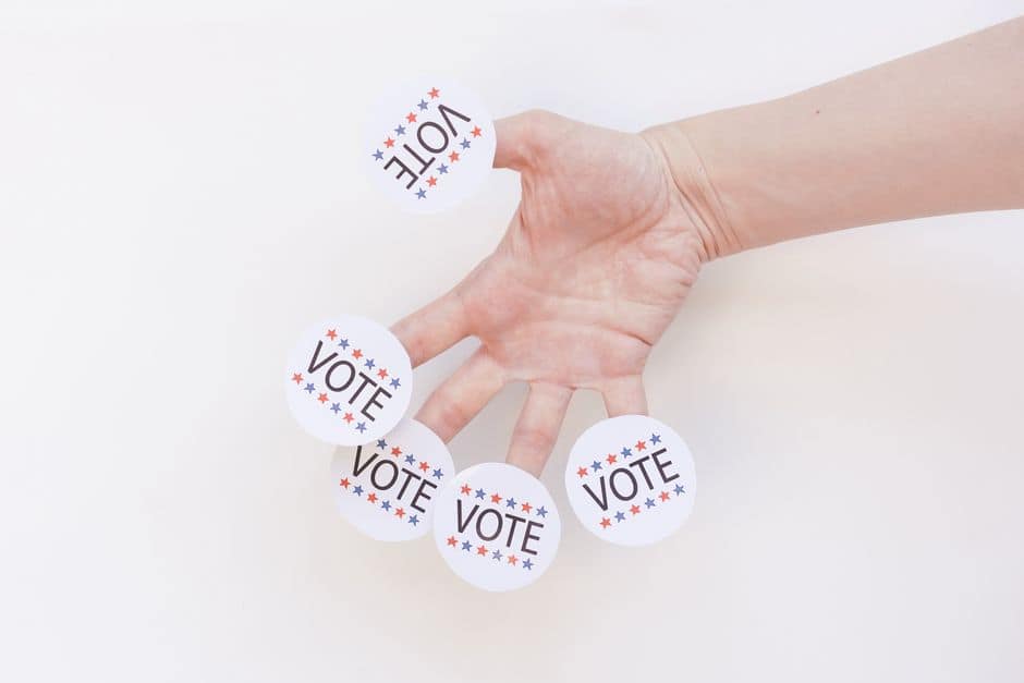 A hand displays several 'Vote' stickers on a white background, symbolizing political participation.