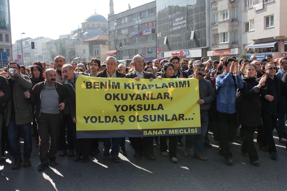 A diverse crowd protesting in an urban area, holding a notable banner.