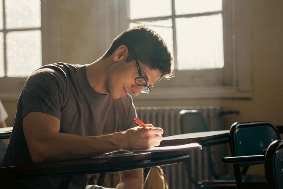 A student concentrating on writing during a class session in a warmly lit university classroom.