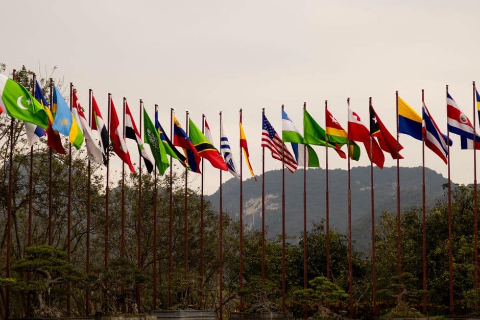 A display of various national flags on flagpoles set against a mountain backdrop, symbolizing global unity.