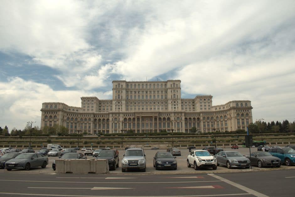 A daytime view of the iconic Palace of the Parliament in Bucharest, Romania, featuring the grand architecture and parked cars.