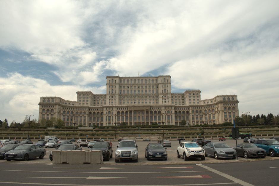 A daytime view of the iconic Palace of the Parliament in Bucharest, Romania, featuring the grand architecture and parked cars.